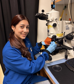 Jazmin in a blue lab coat and gloves smiles while using a microscope in a laboratory.