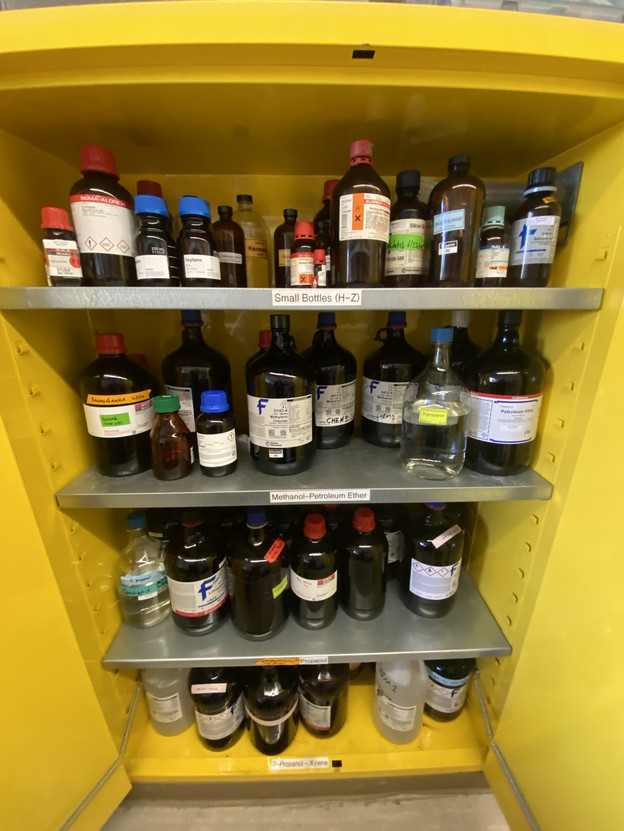 A yellow cabinet with doors open, showing four shelves filled with chemical containers.